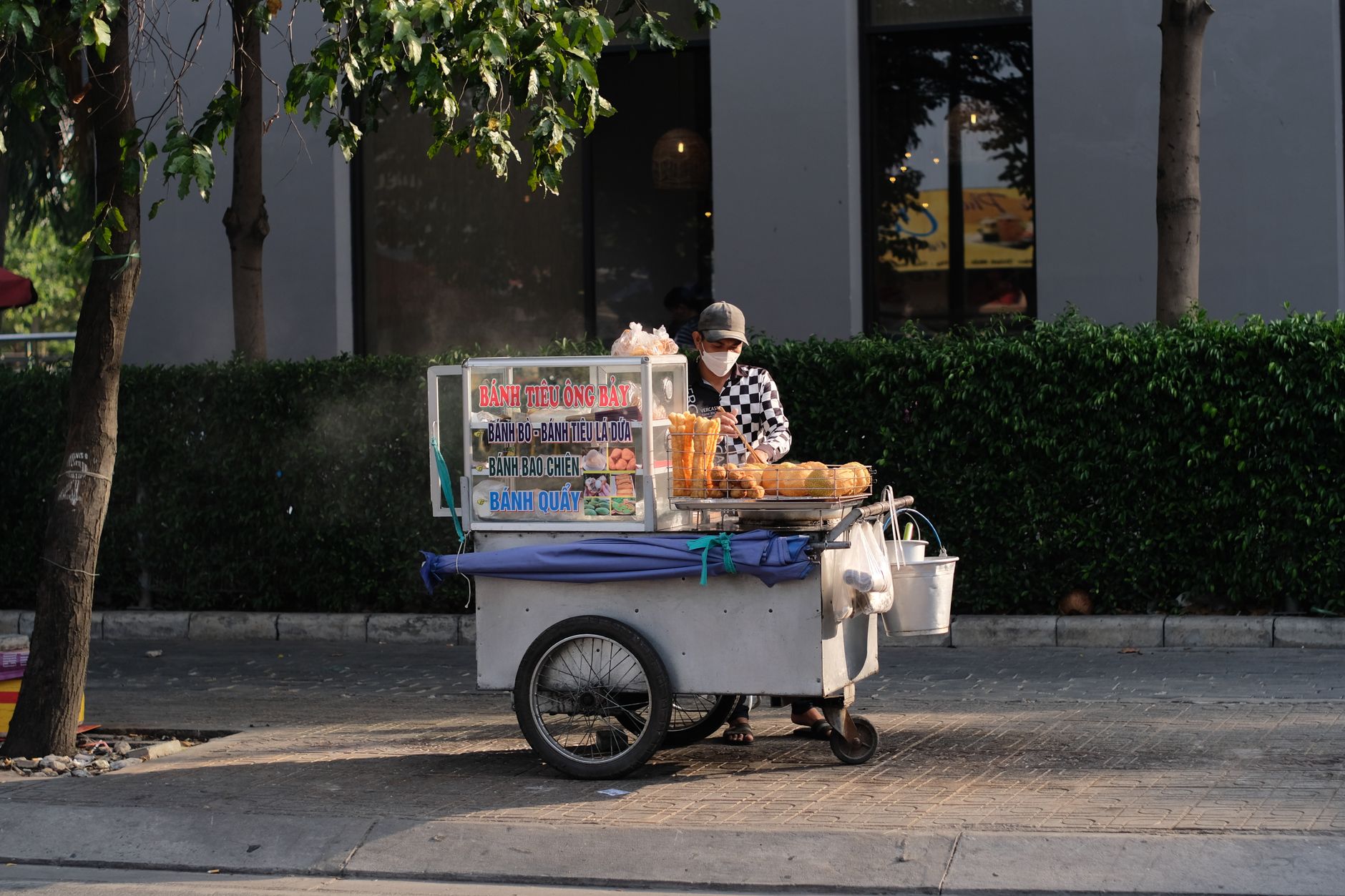Nearby Ideas — A street vendor with a cart selling bánh tiêu and other pastries on a sunny day. (photo: Nguyen Huy)
