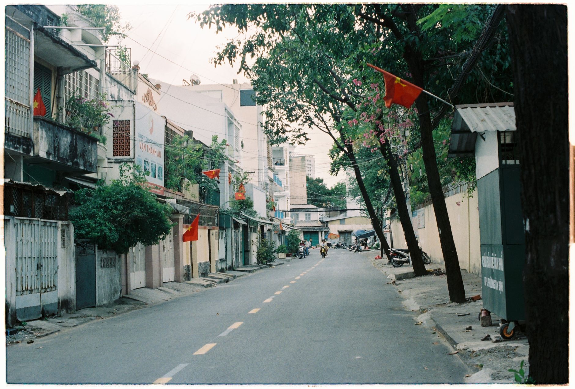 What to Expect on the Ground — Quiet urban street lined with flags and trees, showcasing residential architectu (photo: Thới Nam Cao)