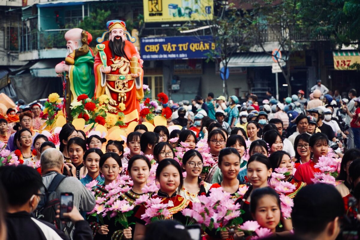 A vibrant street festival parade featuring Asian participants with floral decorations and large statues. — photo: Tuan Vy