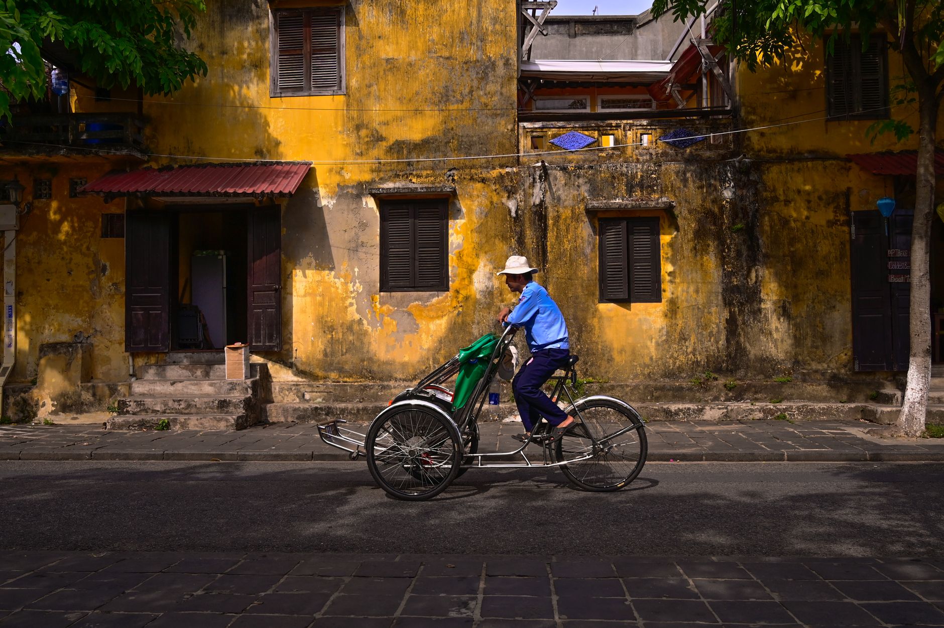 Nearby Ideas — A man rides a tricycle past a weathered yellow wall in an urban setting. (photo: Nguyen Ngoc Tien)